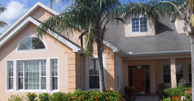 Exterior view of a peach-colored building with white trim, featuring large windows with white blinds, a gray shingled roof, and palm trees in front. There is a covered entrance with an open door and some outdoor chairs visible inside the porch area. Green shrubs and flowering plants are in the foreground under a partly cloudy blue sky.