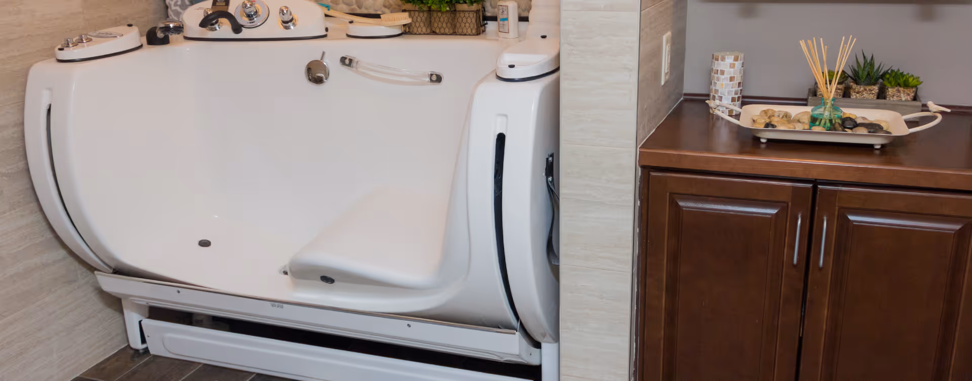 A white walk-in bathtub with built-in seat and multiple control knobs, adjacent to a wooden cabinet with decorative items including a tray with stones and reed diffuser.