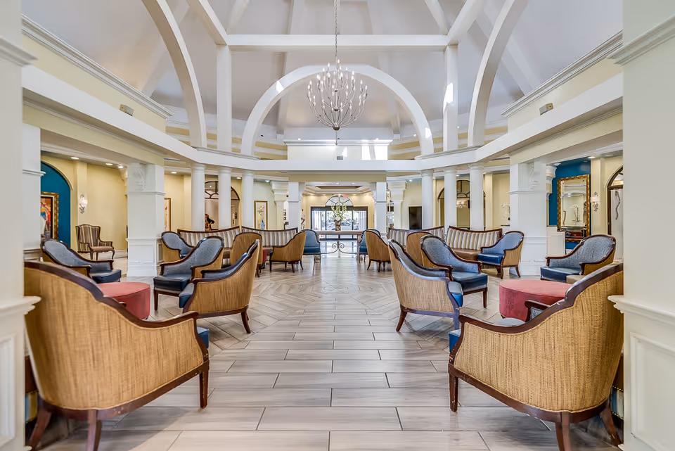 Spacious senior living facility common area with high vaulted ceilings, white arches, and a large chandelier. The room is furnished with multiple sets of wicker and upholstered chairs arranged around small round tables on a tiled floor. The walls are light-colored with decorative columns and artwork, and there is natural light coming through the entrance at the far end.