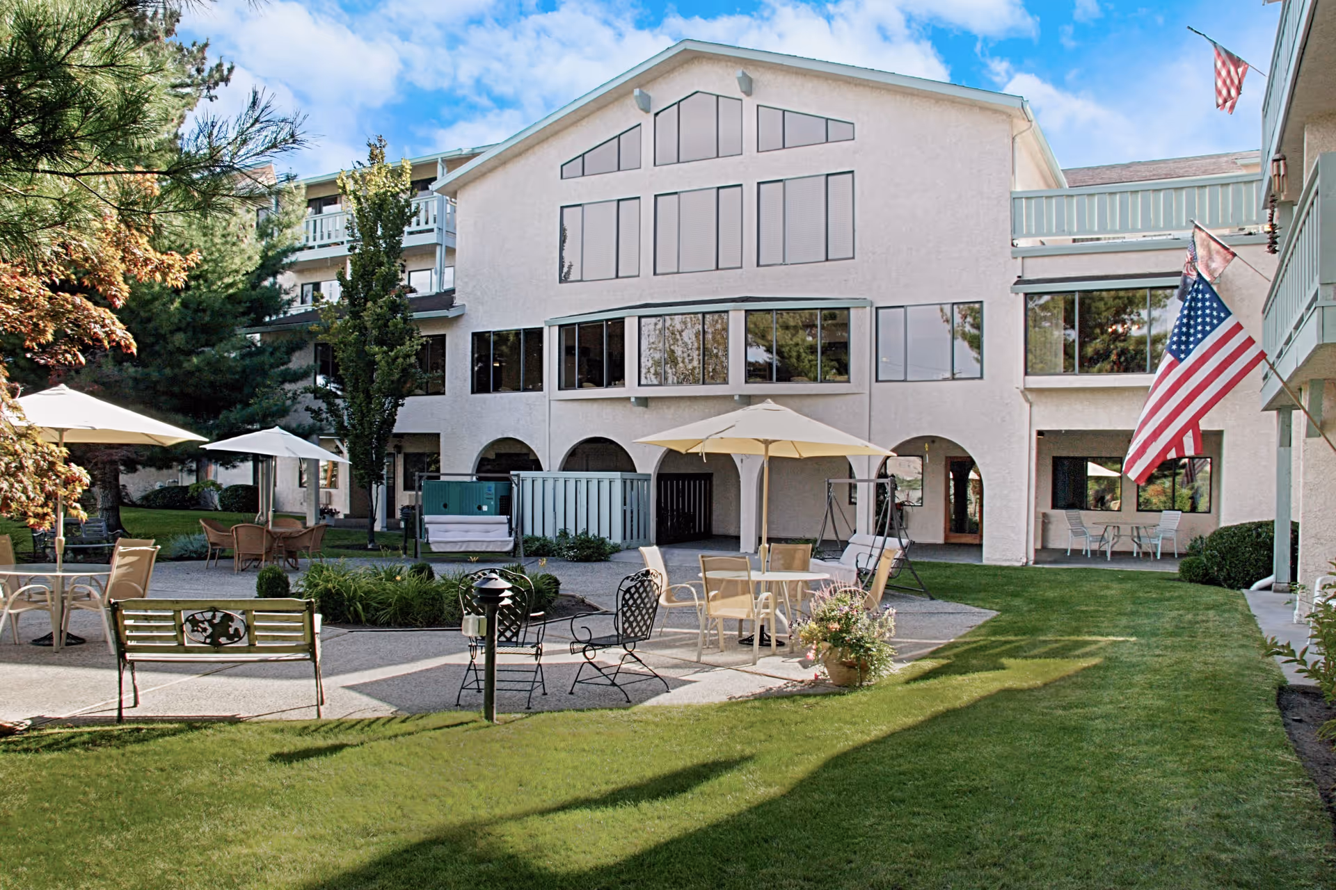 Outdoor courtyard area of a senior living facility with green grass, patio tables with umbrellas, chairs, a bench, and an American flag. The building in the background has large windows and balconies, surrounded by trees and landscaping.