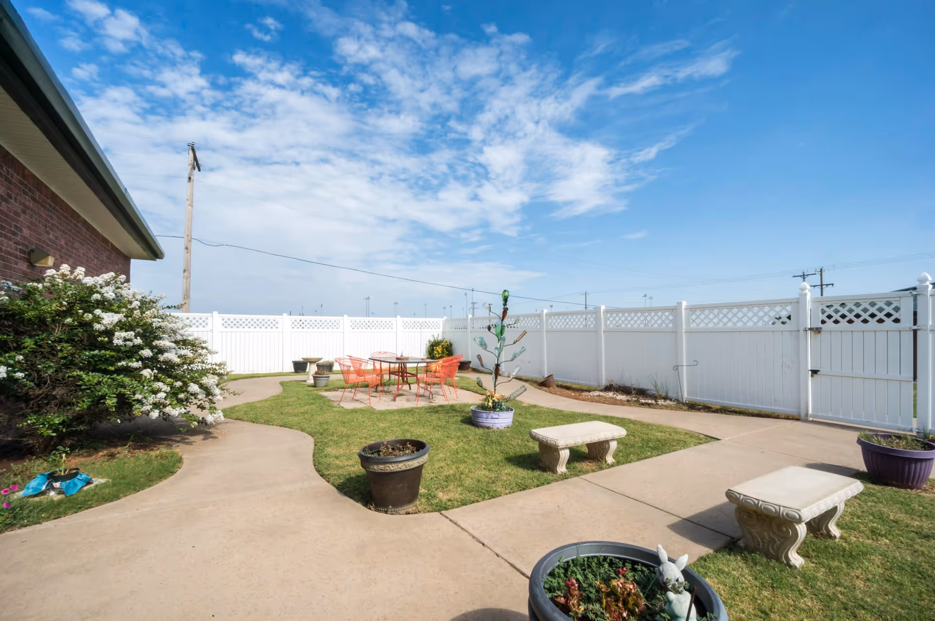 Outdoor patio area at Homestead Senior Living of Elk City featuring a concrete walkway, green grass, potted plants, two stone benches, and a table with orange chairs under a partly cloudy blue sky, enclosed by a white fence.