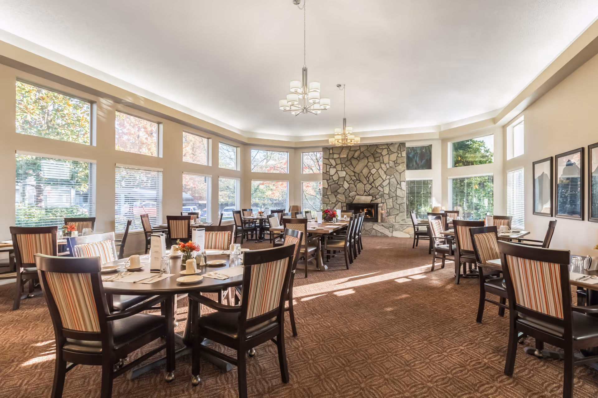 A bright dining room with multiple round and rectangular tables set with cups, plates, and silverware. The room features large windows letting in natural light, a stone fireplace at the far end, and chairs with striped upholstery. The carpet is patterned and the walls are decorated with framed pictures.