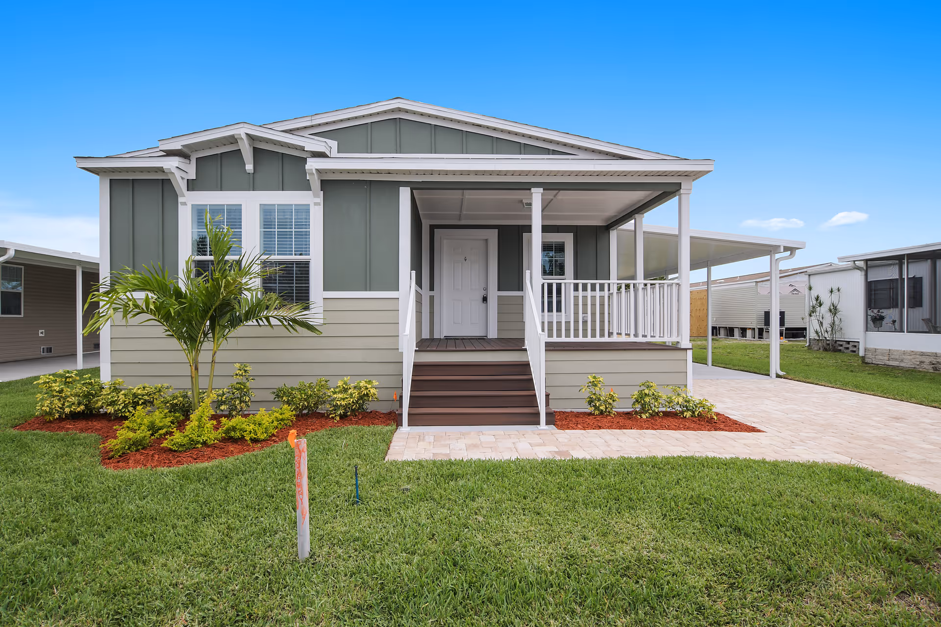 Front view of a single-story manufactured home with a small porch, white railing, and steps leading up to a white front door. The house exterior is painted in green and beige with white trim. There is a small landscaped garden with a palm tree and shrubs in front of the house, and a paved driveway to the right. The sky is clear and blue.