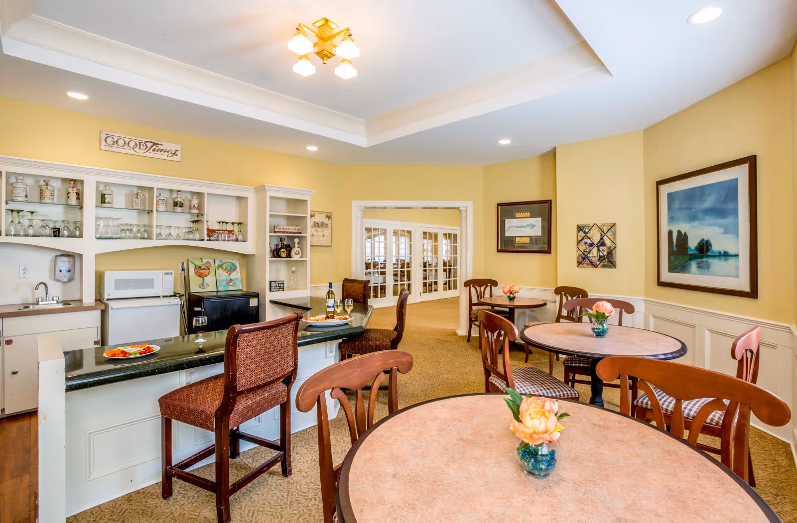 A cozy dining area in a senior living facility with round tables and wooden chairs. Each table has a small vase with a flower. There is a small kitchenette with a countertop, bar stools, a microwave, a mini fridge, and shelves with glassware and decorative items. The walls are painted yellow and decorated with framed artwork and a sign that reads 'Good Times'.