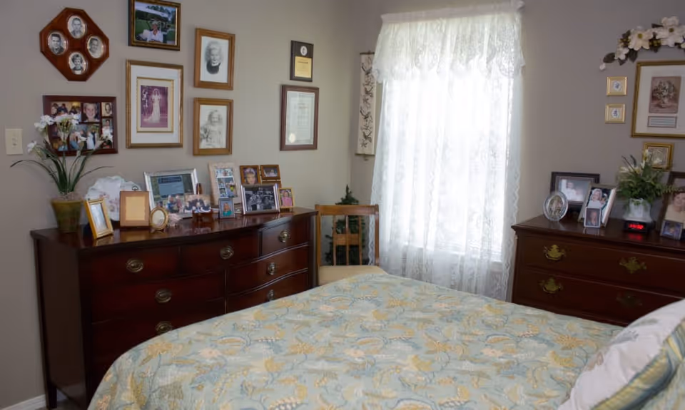 Cozy bedroom with a patterned bedspread, two wooden dressers covered in framed photos and a lace-curtained window.