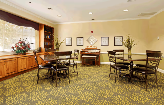 Communal activity room with round tables and chairs, an organ against the back wall, built-in wooden shelving under a window, and a patterned carpet.