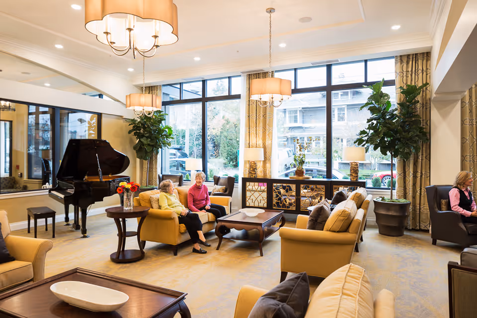A bright and spacious living room area in a senior living facility with large windows letting in natural light. The room features comfortable beige sofas and armchairs arranged around wooden coffee tables. Two elderly women are seated and conversing on one sofa, while another woman sits in a separate armchair. There is a black grand piano in the corner, large potted plants, and elegant hanging light fixtures. The decor includes patterned curtains and decorative lamps on a mirrored console table.