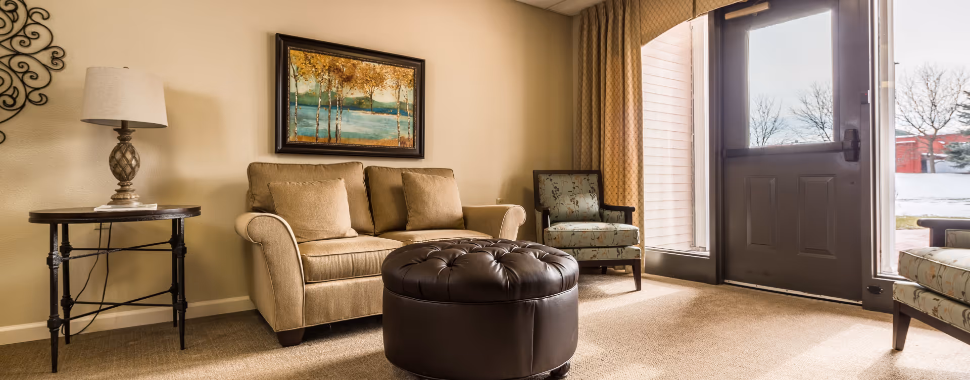 A cozy living room area with a beige loveseat, two patterned armchairs, a round dark brown tufted ottoman, a side table with a lamp, a framed painting of trees on the wall, and a door with large windows letting in natural light.