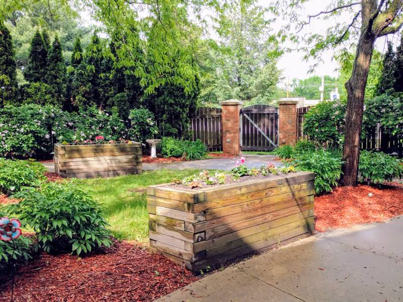 Outdoor garden area with raised wooden planter boxes filled with flowers, surrounded by green shrubs and trees. A brick and wooden gate is visible in the background, along with a paved walkway and mulch-covered ground.