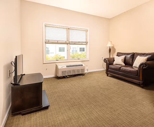 A small living room with beige walls and carpeted floor. There is a dark brown leather couch with two light-colored pillows on the right side, a floor lamp behind it, and a flat-screen TV on a dark wooden stand on the left. A window with blinds is centered on the back wall above a wall-mounted air conditioning unit.