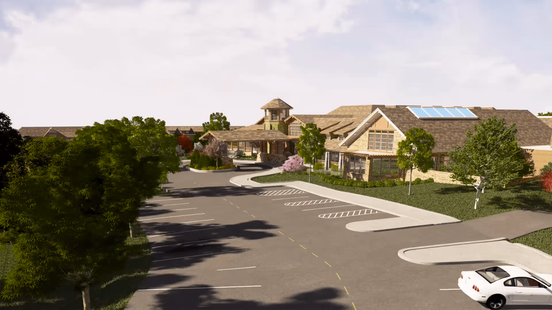 Exterior view of a senior living facility with a large parking lot, trees, and landscaped greenery. The building has a pitched roof with skylights and a covered entrance area.