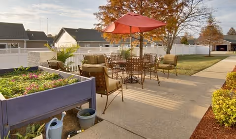 Outdoor patio area with metal chairs and tables, one table shaded by a red umbrella. Raised garden beds with plants and flowers are visible on the left side. The area is surrounded by a white fence, with houses and trees in the background under a partly cloudy sky.