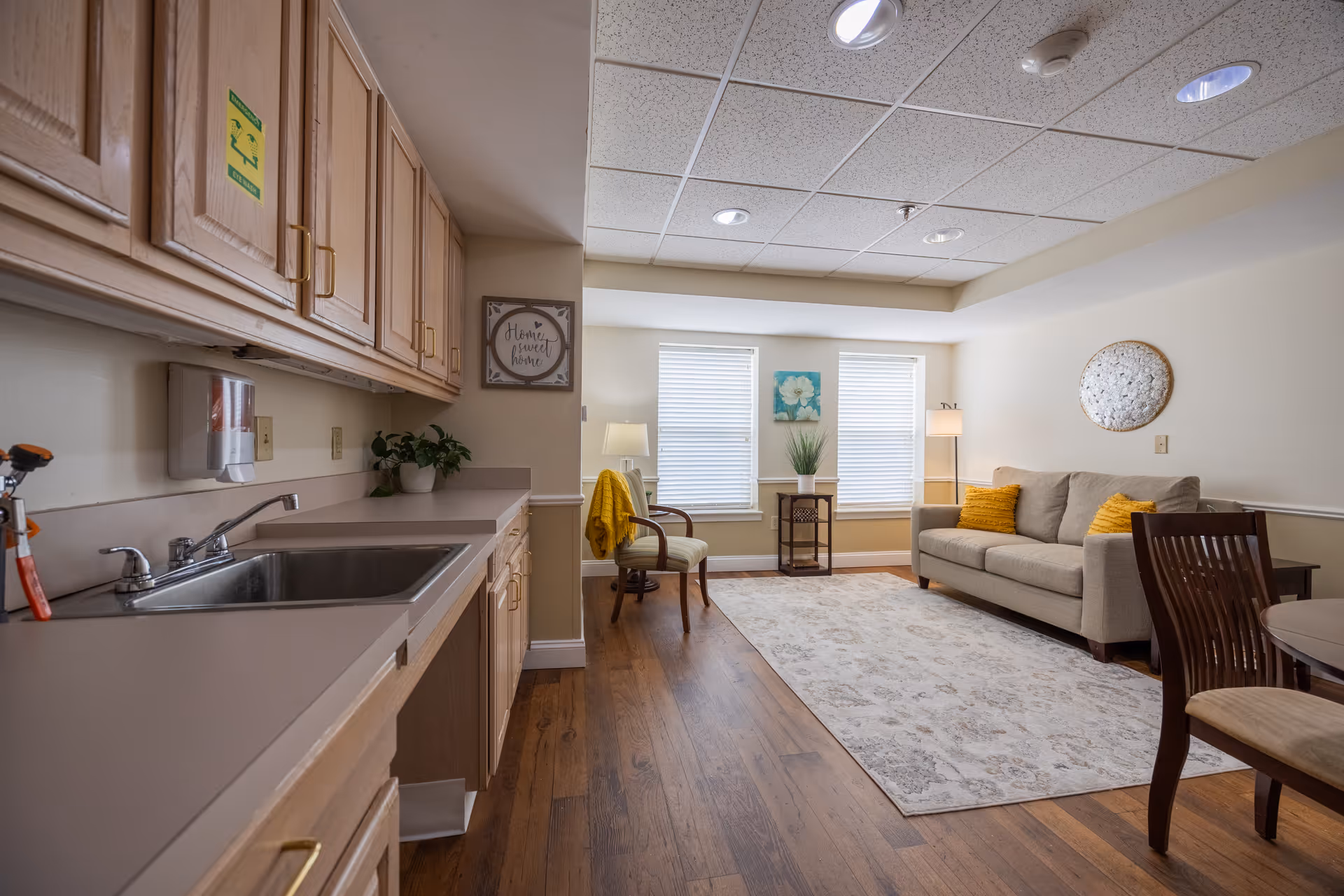 Interior view of a senior living facility room with a kitchenette on the left featuring a sink, cabinets, and countertop. The room extends to a sitting area with a beige sofa adorned with yellow pillows, two chairs, a small table with a plant, and two windows with blinds. The floor is wooden with a large area rug, and the walls are decorated with framed art and a round wall piece.