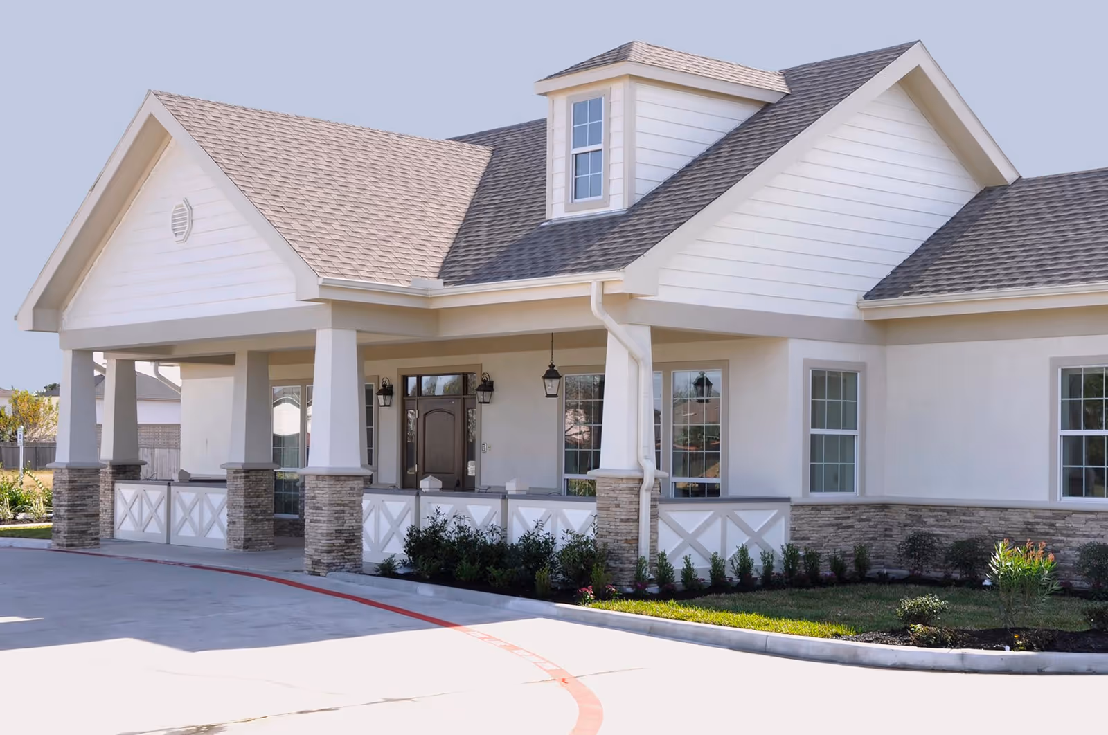 Exterior view of a single-story building with a covered entrance supported by stone and white pillars, featuring a gabled roof with gray shingles, white siding, and multiple windows. There is a small landscaped area with shrubs and grass in front of the building.