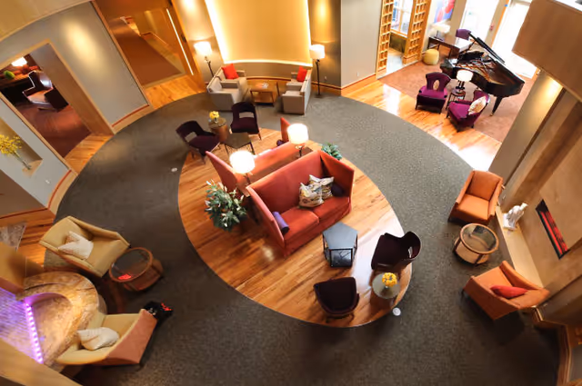 A spacious and well-lit living room area in a senior living facility with various seating arrangements including armchairs, sofas, and small tables. The room features a circular carpet design on a wooden floor, a fireplace, and a grand piano near large windows letting in natural light.