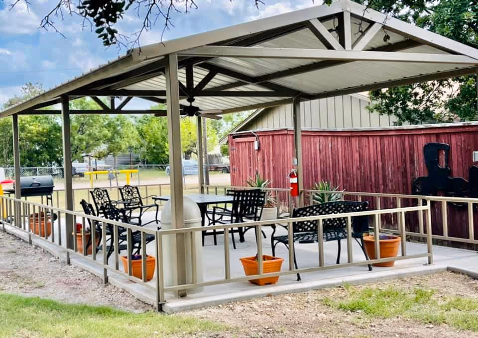Outdoor covered patio area with metal roof and ceiling fan, featuring black metal chairs and tables, several orange plant pots, a grill, and a red wooden fence in the background.