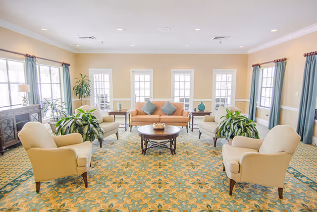 A bright and spacious living room with beige walls and large windows letting in natural light. The room features a patterned carpet in shades of green and yellow, a central round wooden coffee table, a peach-colored sofa with three blue cushions, and four beige armchairs arranged around the table. There are green plants placed near the windows and side tables with decorative items.