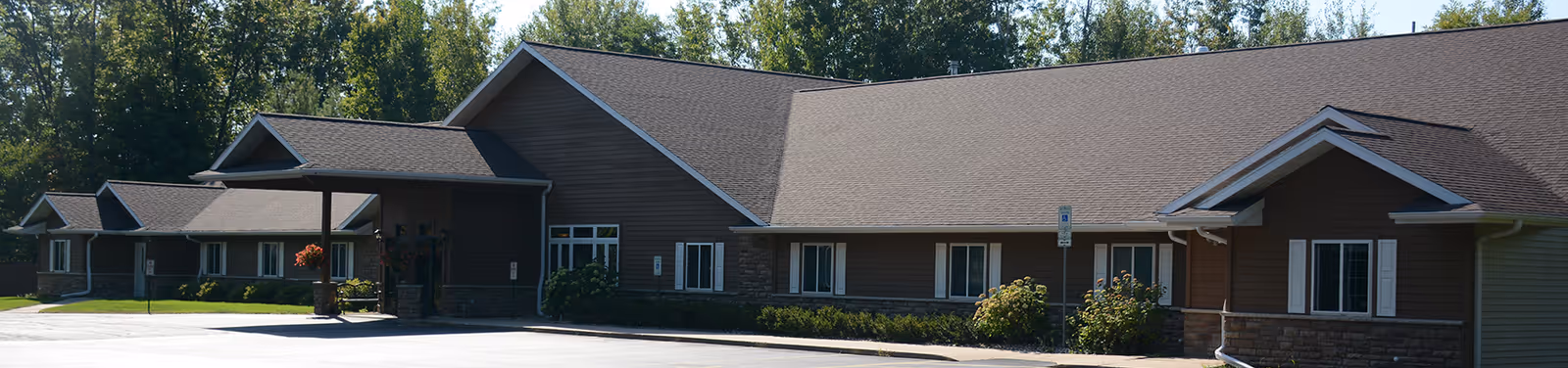 Exterior front view of a single-story assisted living facility building with brown siding and a covered entrance, surrounded by greenery and trees in the background.