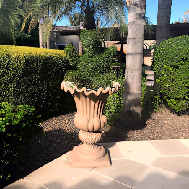 Outdoor garden area with a large decorative planter filled with green shrubs, surrounded by neatly trimmed hedges and trees, with a building partially visible in the background under a clear blue sky.