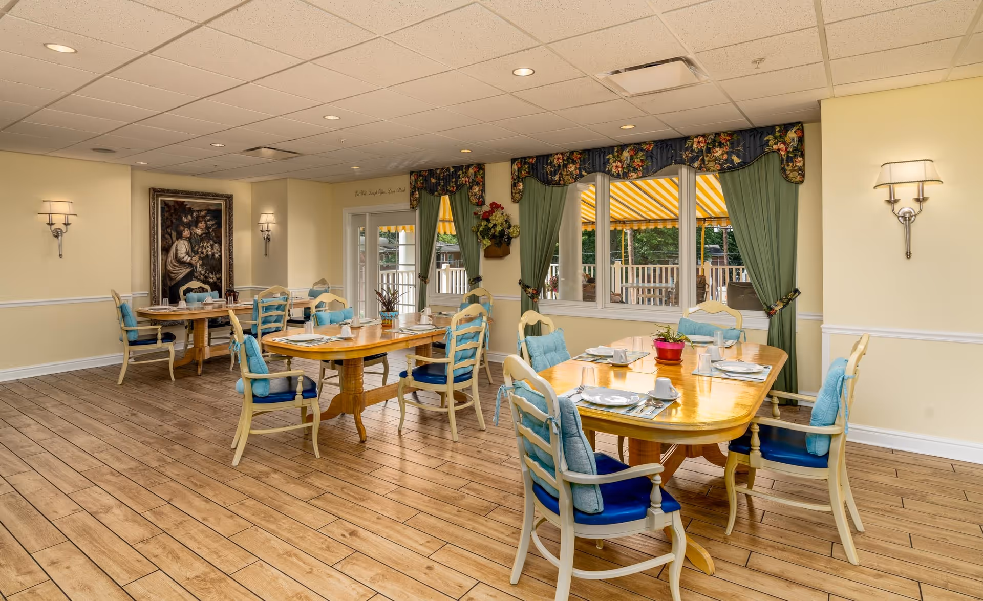A bright dining room with wooden floors and cream-colored walls. There are two wooden dining tables, each surrounded by chairs with blue cushions and light-colored frames. The tables are set with plates, cups, and silverware. Large windows with green curtains and floral valances let in natural light and offer a view of an outdoor patio with a yellow striped awning. Wall sconces provide additional lighting, and a framed painting hangs on one wall.