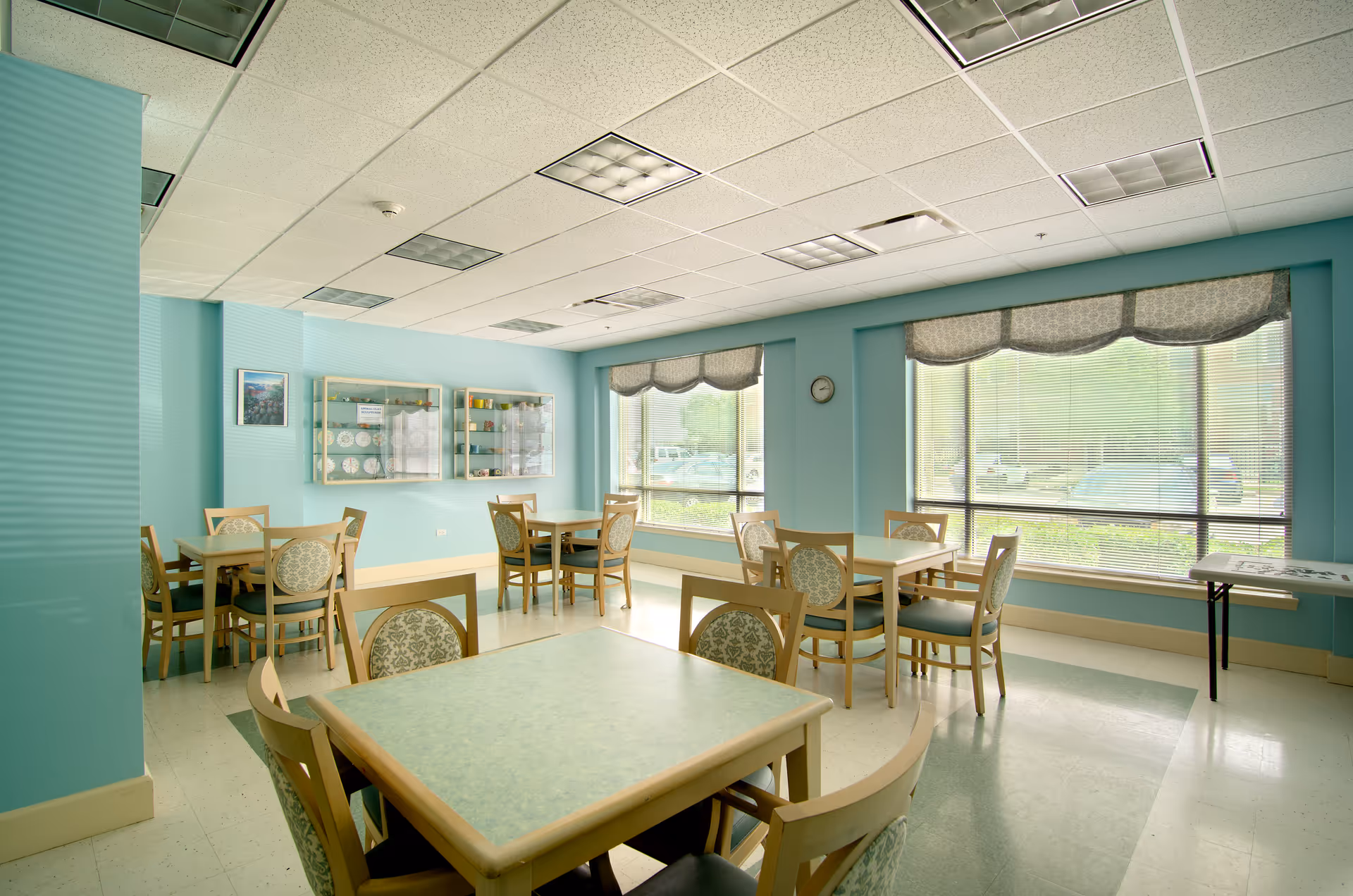 A bright dining room with multiple square tables and wooden chairs with patterned cushions. The walls are painted light blue, and there are large windows with blinds and valances letting in natural light. A clock is mounted on the wall, and display cabinets with plates and cups are visible. The floor is tiled, and the ceiling has recessed lighting panels.