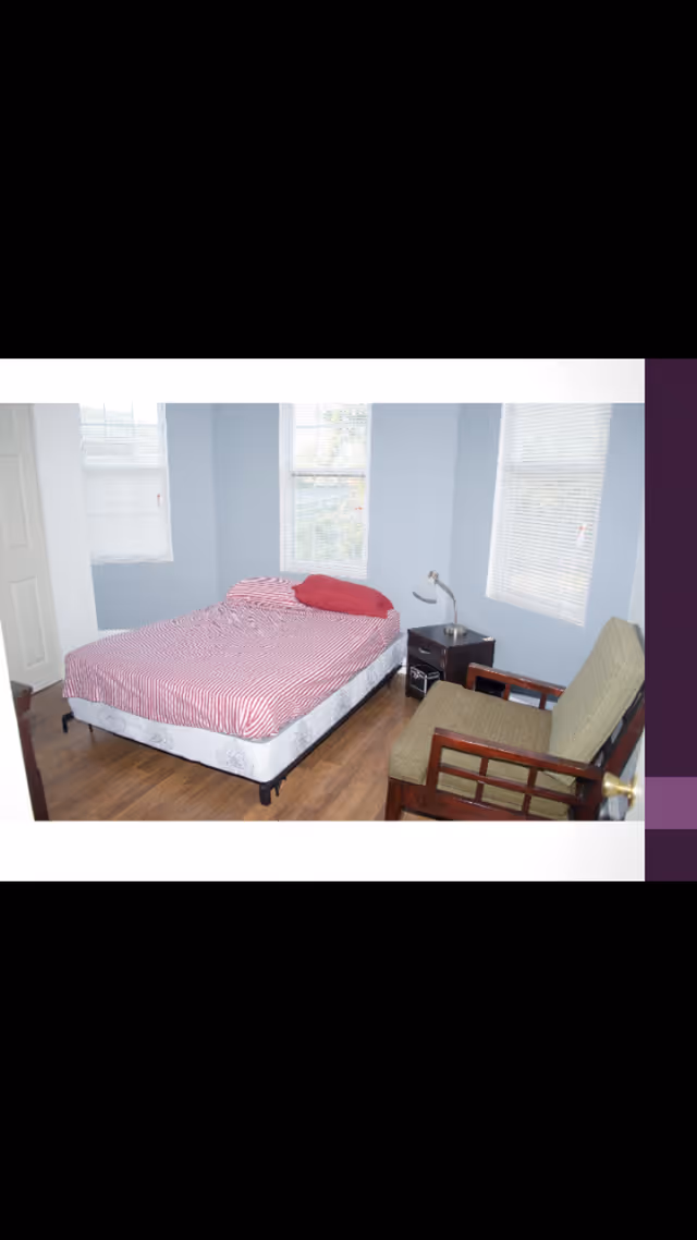 A bedroom with light blue walls and three windows with blinds. The room contains a bed with a red and white striped bedspread and a red pillow, a small dark nightstand with a silver lamp, and a wooden armchair with green cushions. The floor is wooden.