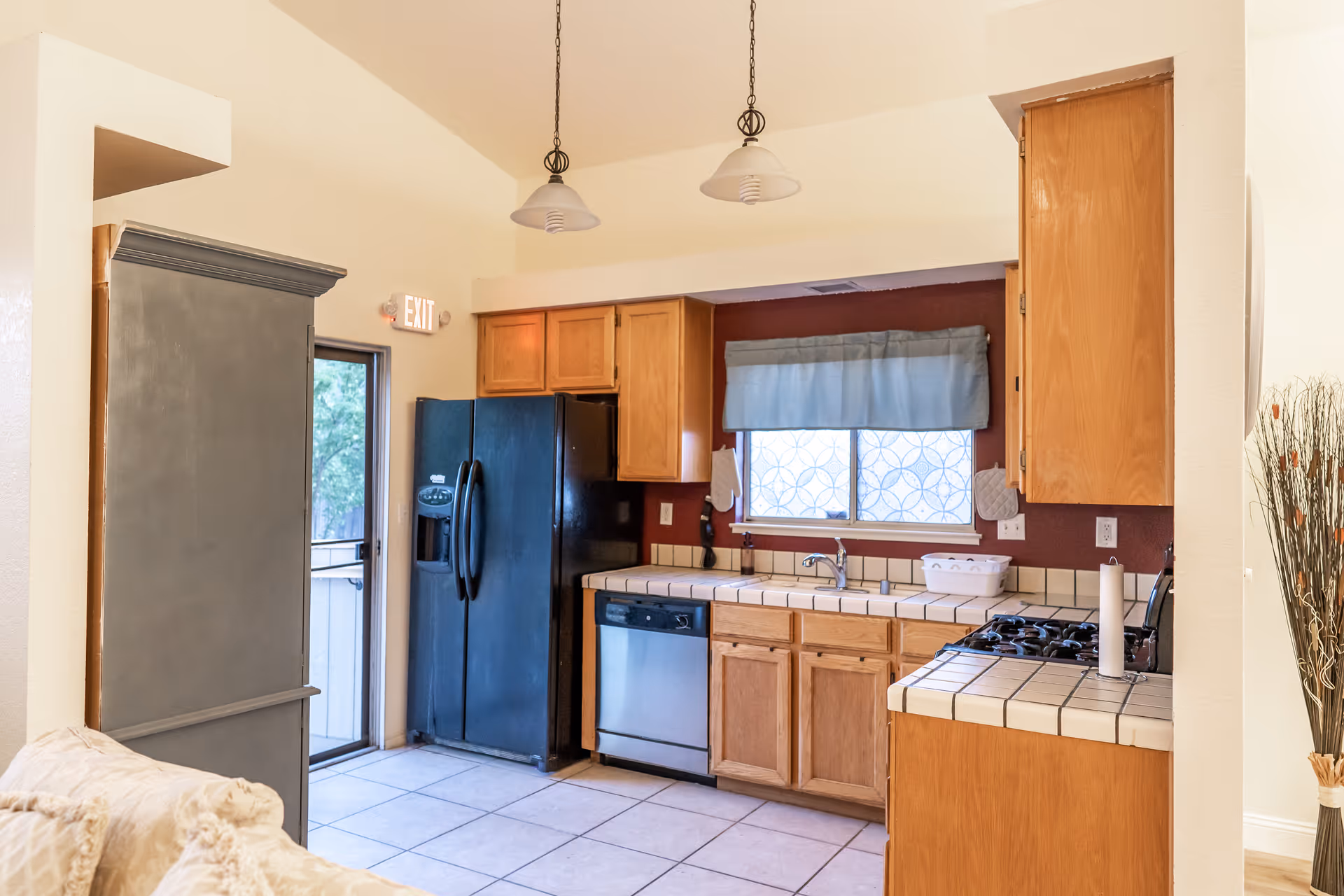 A kitchen area with wooden cabinets, a black refrigerator, a dishwasher, a gas stove, and a tiled countertop. There is a window with a blue valance above the sink, two hanging pendant lights, and a door leading outside. Part of a beige couch and a tall gray cabinet are visible on the left side.