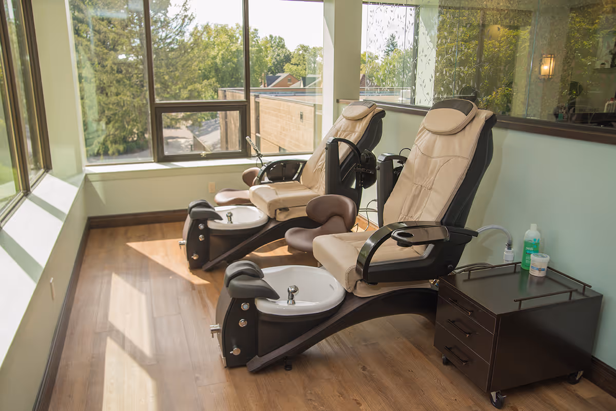 Two beige pedicure spa chairs with foot basins along a sunlit windowed wall in a salon area.