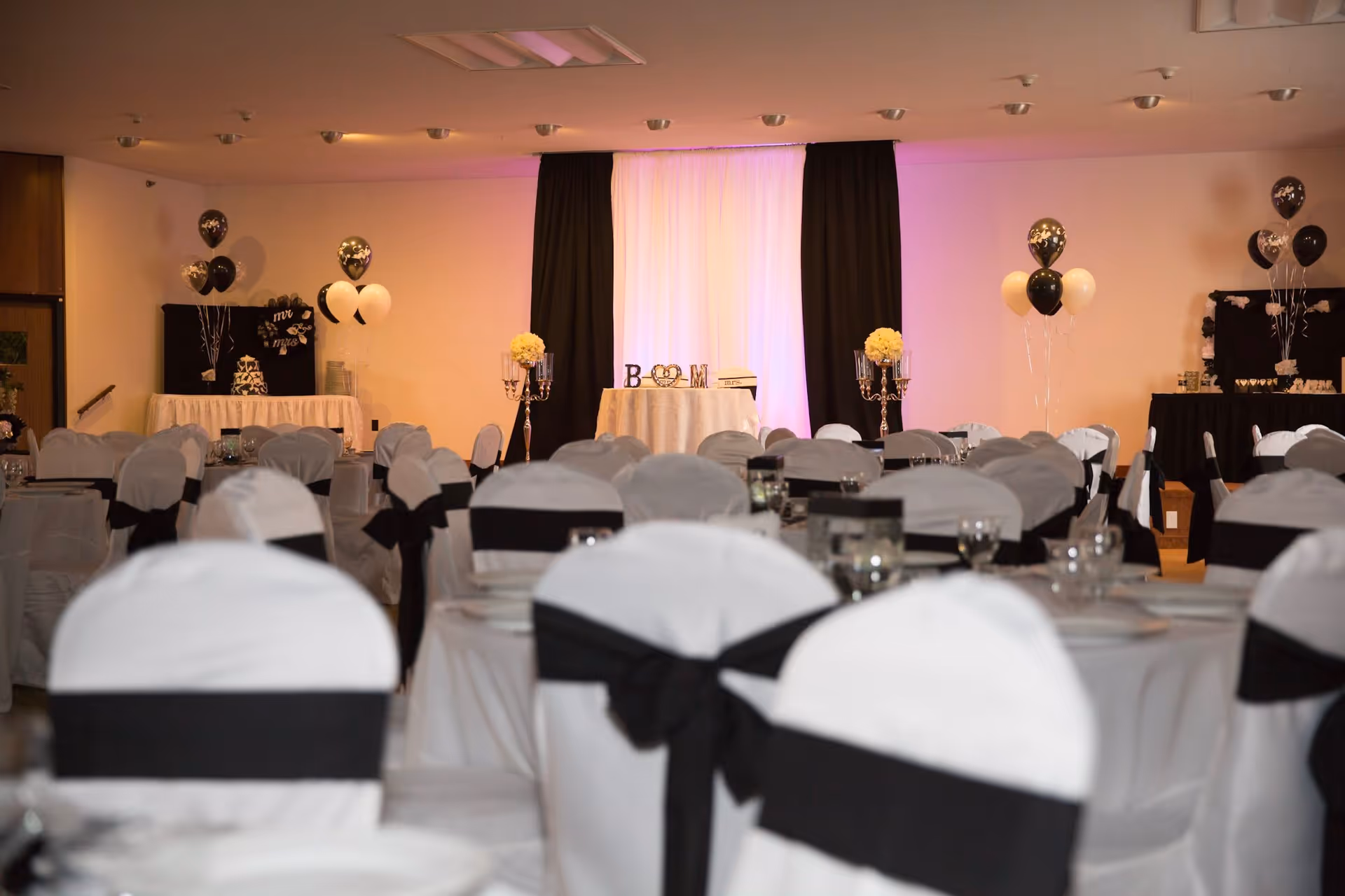 A decorated event room with round tables covered in white tablecloths and chairs with white covers and black sashes. The room features black and white balloons, floral centerpieces, and a backdrop with draped white and black curtains illuminated with soft purple lighting.