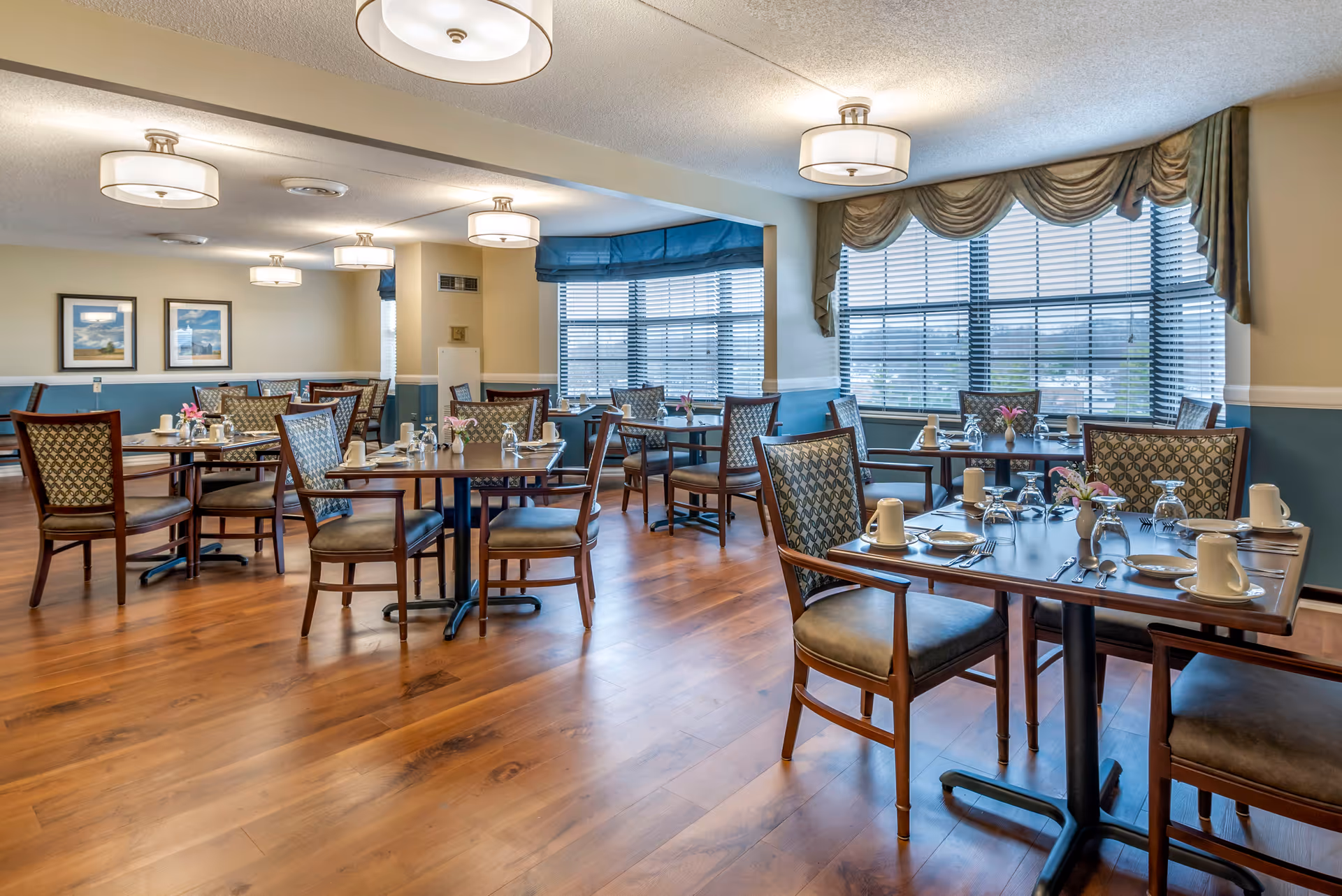 A bright dining room with wooden floors, multiple tables set with plates, cups, utensils, and small flower vases. The room has large windows with blinds and draped curtains, and ceiling lights providing warm illumination.