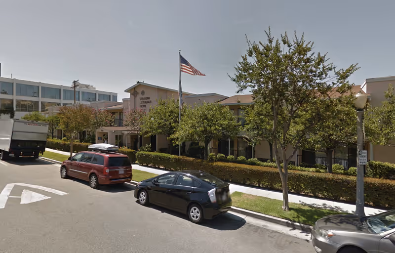 Exterior view of Solheim Lutheran Home, a senior living facility, showing a two-story building with an American flag on a flagpole in front. Several cars are parked along the street, and there are trees and bushes lining the sidewalk.