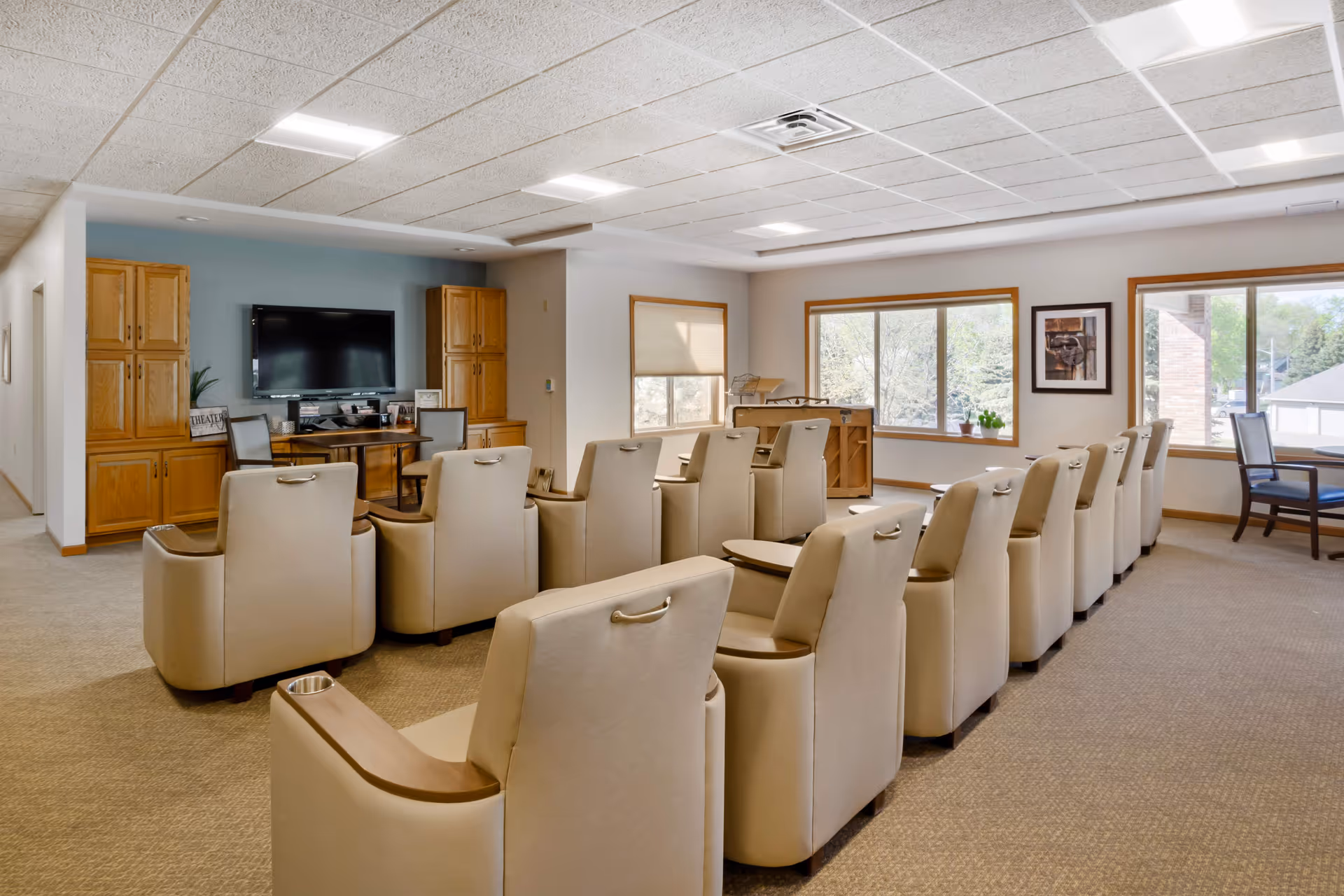 A spacious senior living common room with two rows of beige recliner chairs facing a wall-mounted TV and large windows.