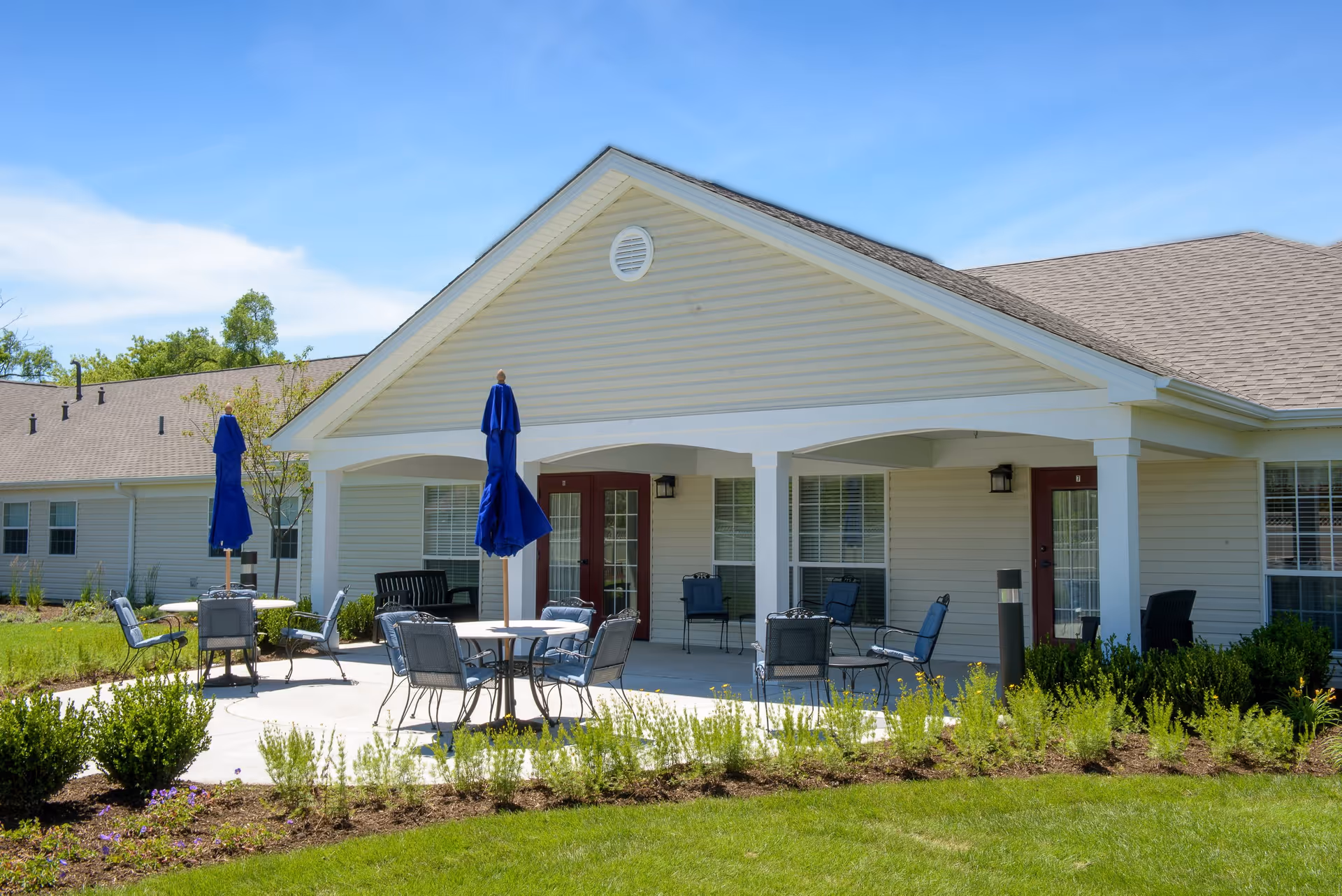 Outdoor patio area of a senior living facility with several metal chairs and tables, some with blue umbrellas. The patio is adjacent to a beige building with white trim and red doors, surrounded by green grass and landscaped plants under a clear blue sky.