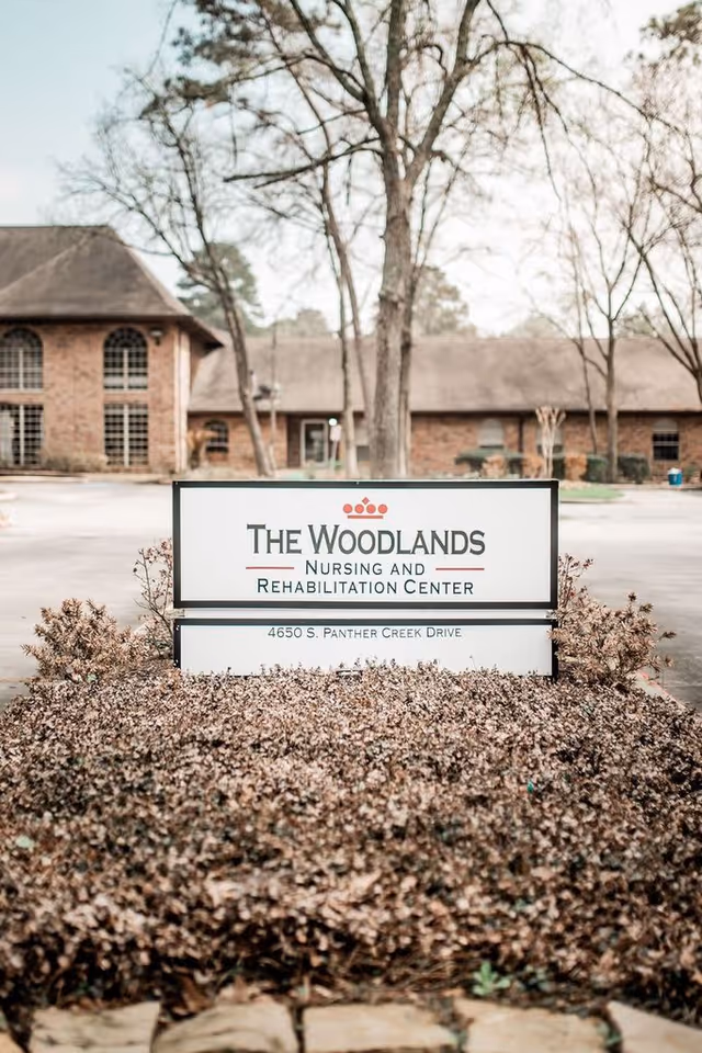 Outdoor view of The Woodlands Nursing and Rehabilitation Center sign with the building and leafless trees in the background.
