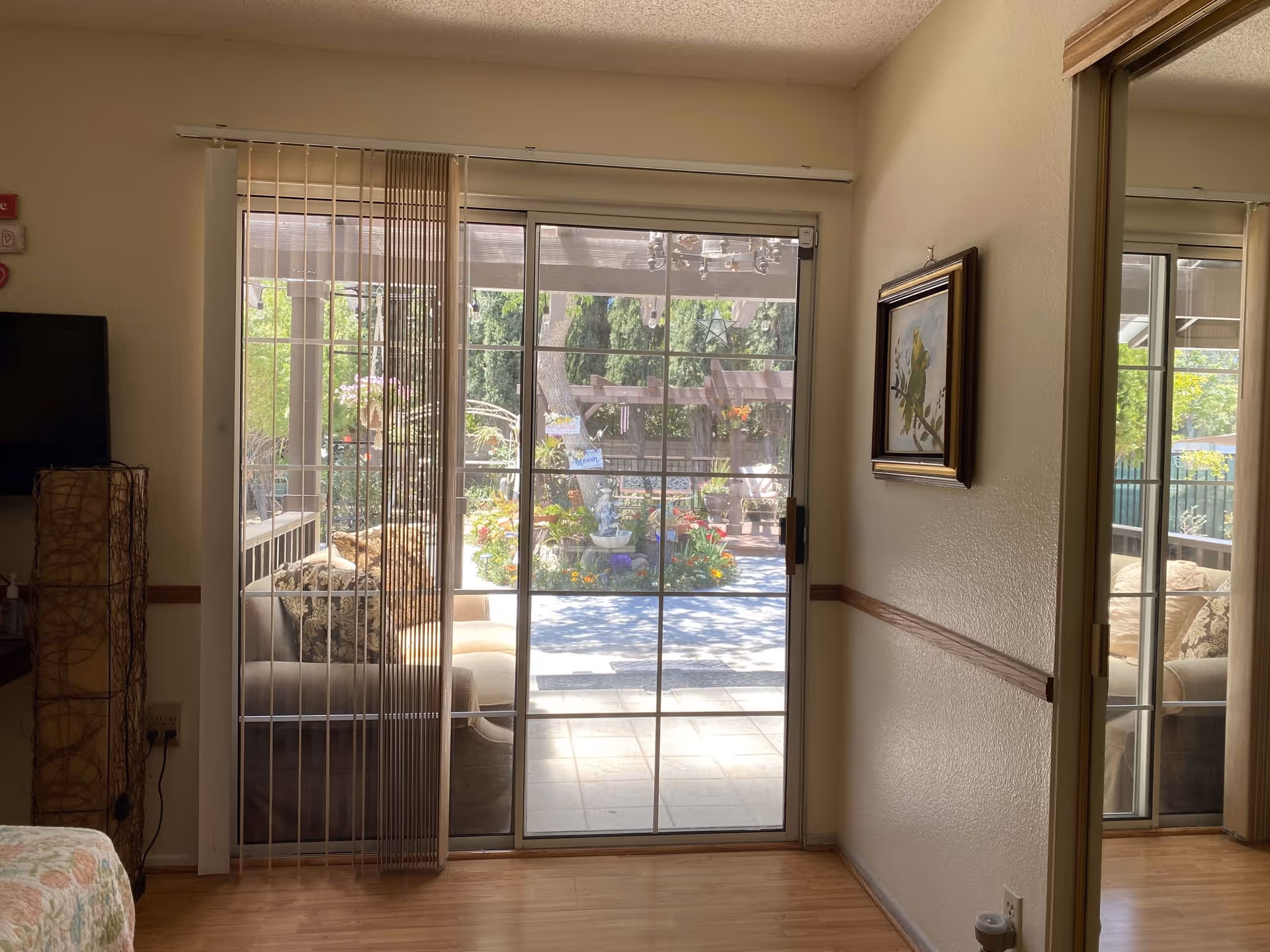 Interior view of a room with wooden flooring, a sliding glass door leading to a garden patio with plants and flowers. There is a couch partially visible on the left side, a tall lamp with a woven design, a wall-mounted TV, and a framed painting of birds on the right wall. The patio outside has a pergola and various colorful plants.