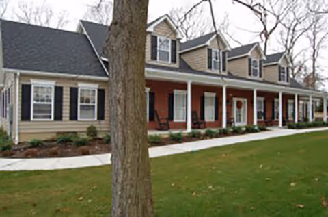 Front exterior of a residential-style building with a covered porch, dormer windows, and a tree in the foreground.