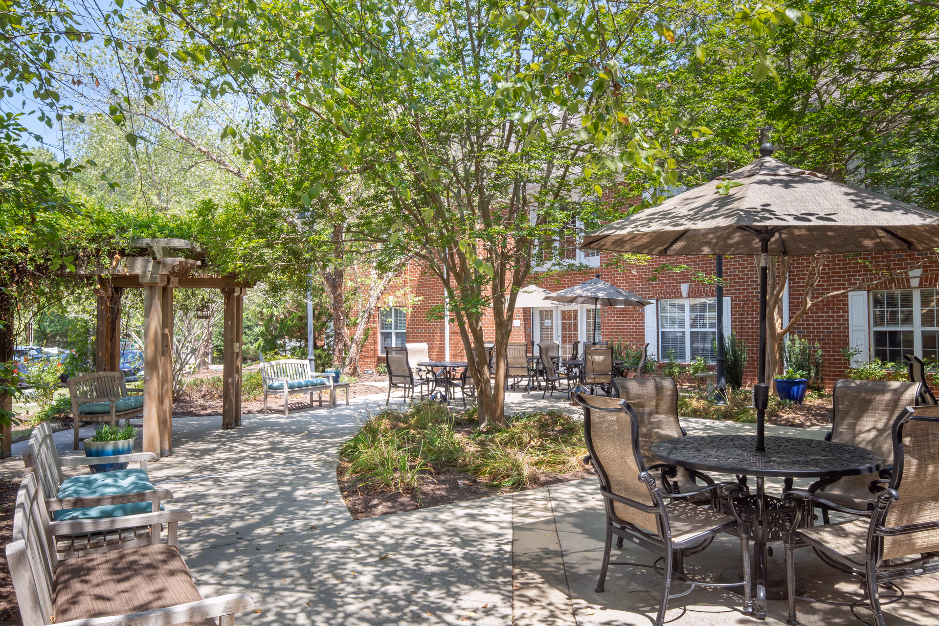 Shaded outdoor courtyard with patio tables and umbrellas, benches, a pergola, and a brick building in the background.