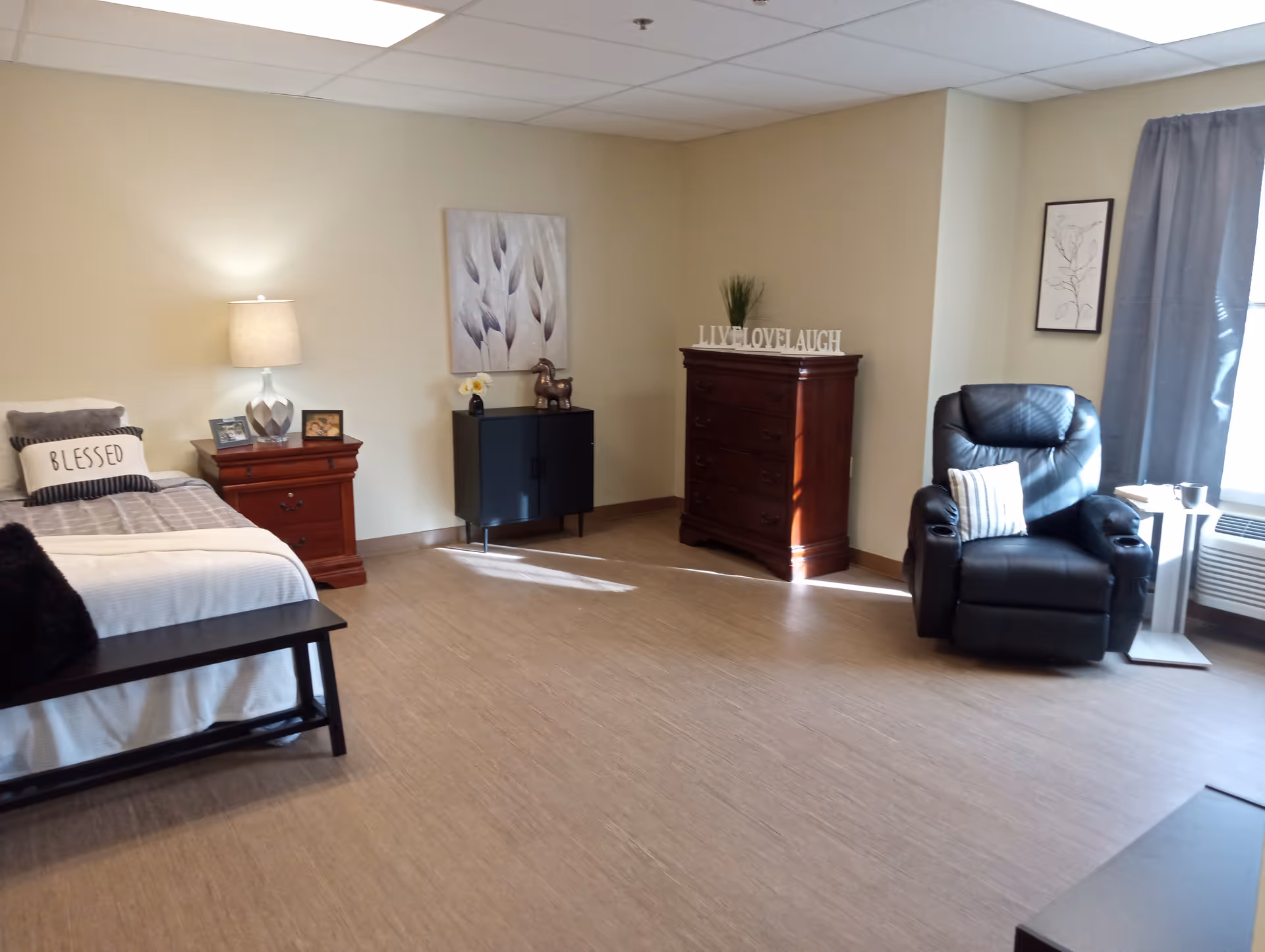 A well-lit bedroom with a single bed on the left side, a wooden nightstand with a lamp and picture frames, a black cabinet with decorative items, a wooden chest of drawers with a 'LIVE LOVE LAUGH' sign on top, a black leather recliner chair with a striped pillow near a window with gray curtains, and light-colored walls and flooring.