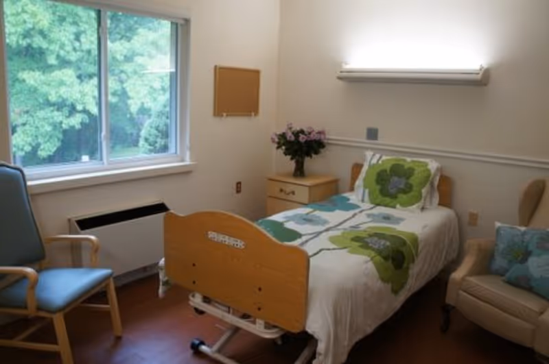 Single-occupancy senior room with a hospital-style bed dressed in floral bedding, a nightstand with flowers, chairs, and a window overlooking trees.