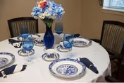 A round dining table set for four with blue-and-white china, blue glassware, navy napkins, and a blue vase of flowers in a dining room.