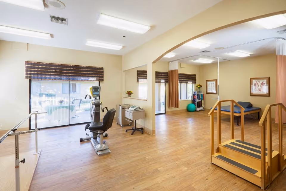 A bright rehabilitation room with wooden flooring, featuring exercise equipment including a stationary bike, parallel bars for walking practice, a set of wooden stairs with handrails, and a therapy table. Large windows with brown blinds allow natural light to enter, and there are anatomical charts on the walls along with a green exercise ball and a rack of therapy supplies.