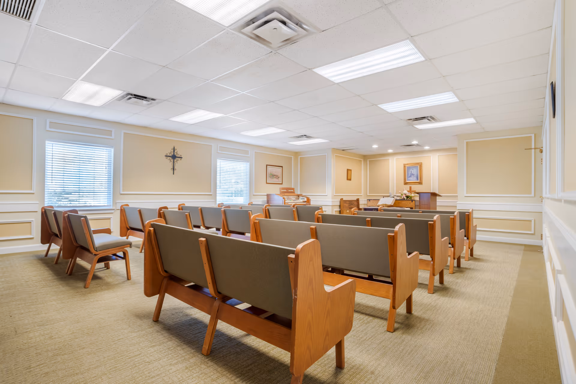 Interior view of a chapel or small worship room with rows of wooden pews with gray cushions, beige walls with white trim, two windows with blinds, a cross on the wall, and framed pictures. The room is well-lit with ceiling lights.