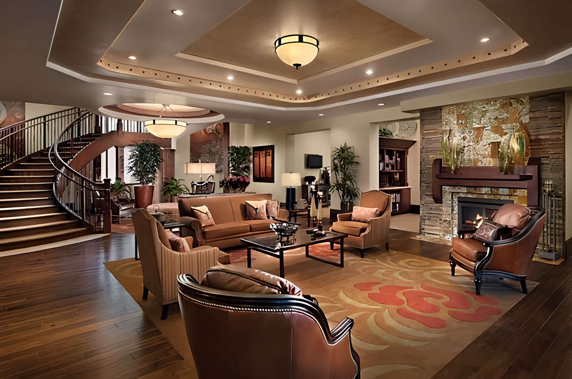 A spacious and elegant living room in an assisted living facility featuring a large area rug with floral patterns, a fireplace with decorative stonework, multiple comfortable armchairs and sofas arranged around a central coffee table, potted plants, warm lighting from ceiling fixtures, and a curved wooden staircase in the background.