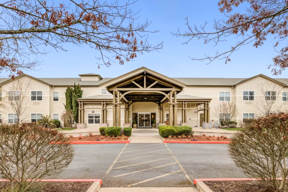 Front exterior view of a two-story senior living facility building with a covered entrance supported by wooden beams, surrounded by trimmed bushes and leafless trees under a clear sky.