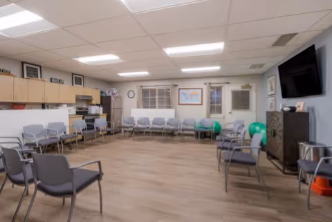 Spacious senior living common room with chairs arranged around the perimeter, a kitchenette, wall-mounted TV, and exercise balls.