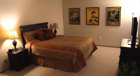 A bedroom in a senior facility with a large bed, lamps, nightstands, and wall posters.