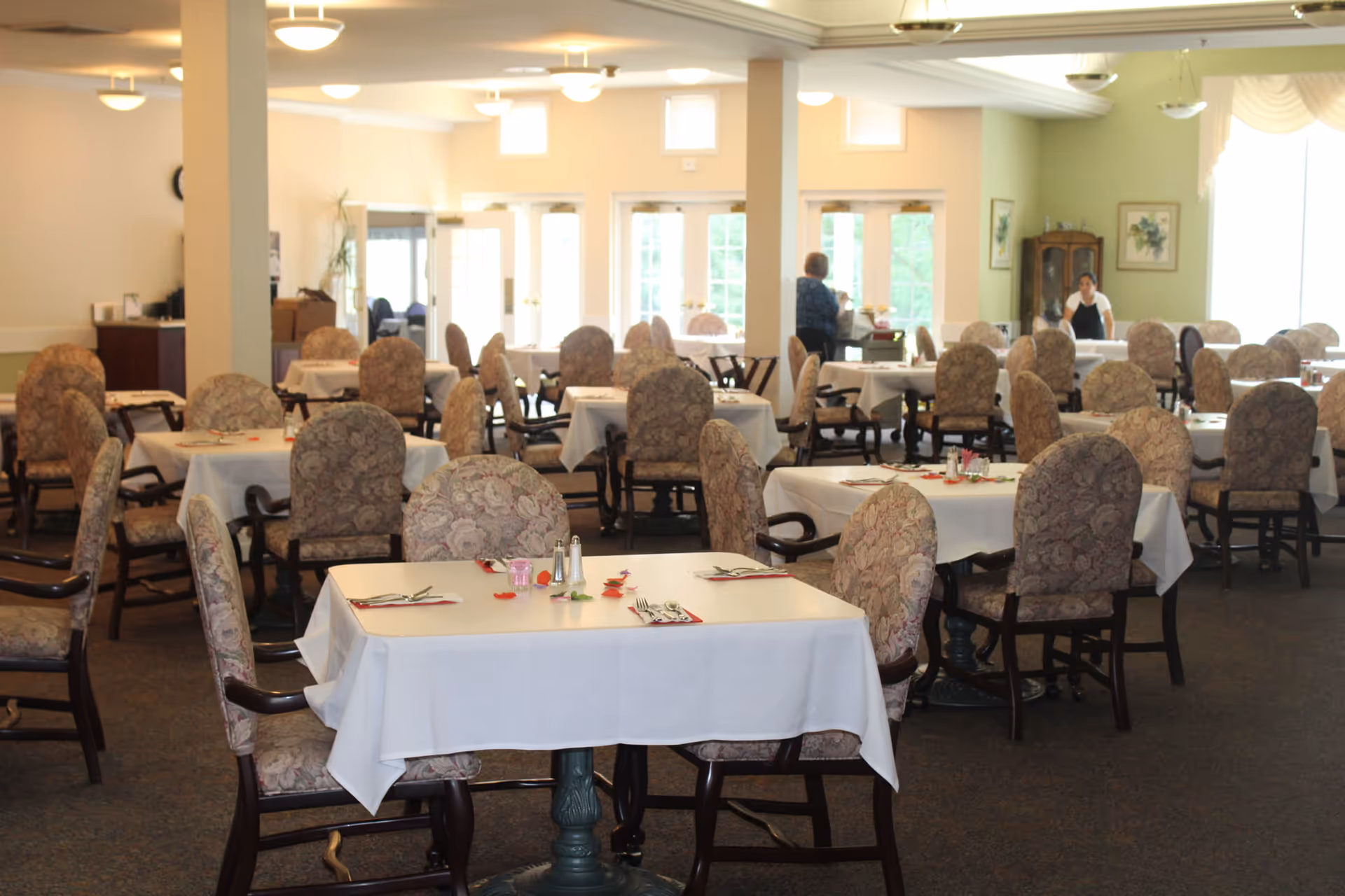 A spacious dining room with multiple tables covered in white tablecloths, each set with utensils and napkins. The chairs have floral upholstery and wooden armrests. Two people are visible in the background near the windows and a cabinet. The room is well-lit with ceiling lights and natural light from large windows.