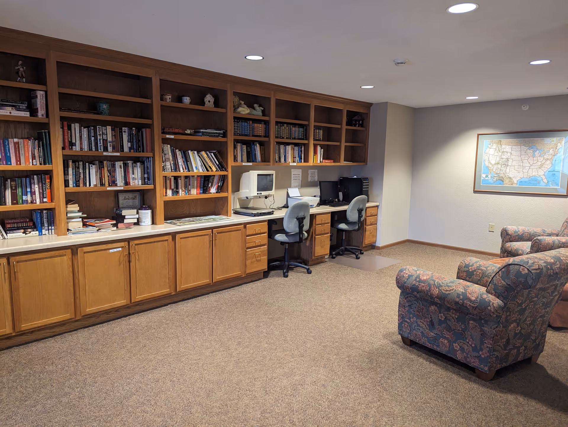 A cozy room with built-in wooden bookshelves filled with books and decorative items. Below the shelves are cabinets and a countertop with two computer workstations, each with an office chair. The room has carpeted flooring and two floral upholstered armchairs. A framed map of the United States hangs on the wall.