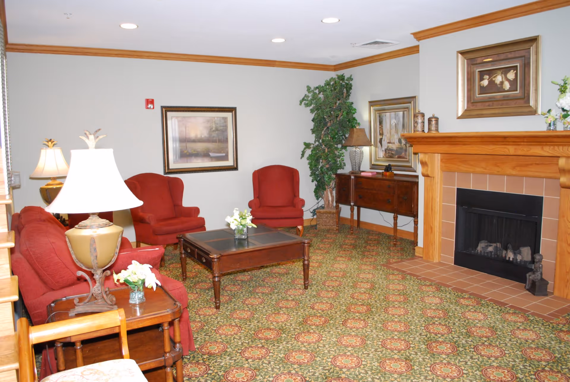 A cozy living room with red upholstered chairs and a matching sofa arranged around a wooden coffee table. The room features a patterned carpet, a wooden fireplace with a tiled hearth, framed artwork on the walls, two table lamps, a potted plant, and a wooden sideboard with decorative items.