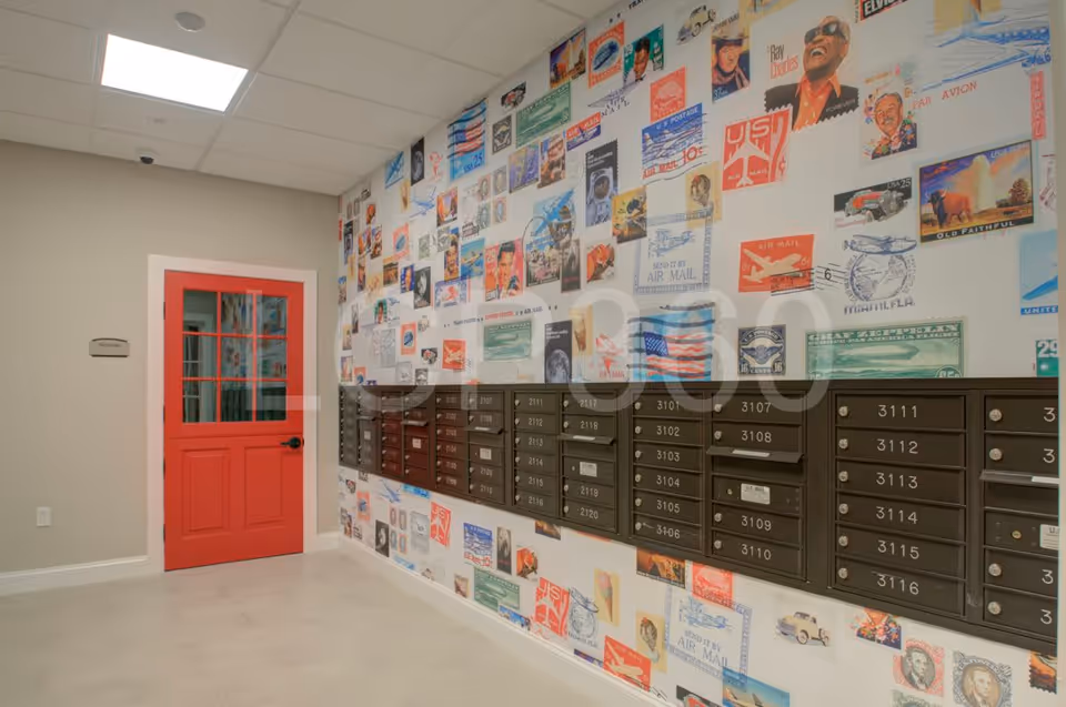 Interior hallway area with a bright red door on the left and a wall of mailboxes on the right. The wall behind the mailboxes is decorated with a colorful collage of vintage postage stamp designs.